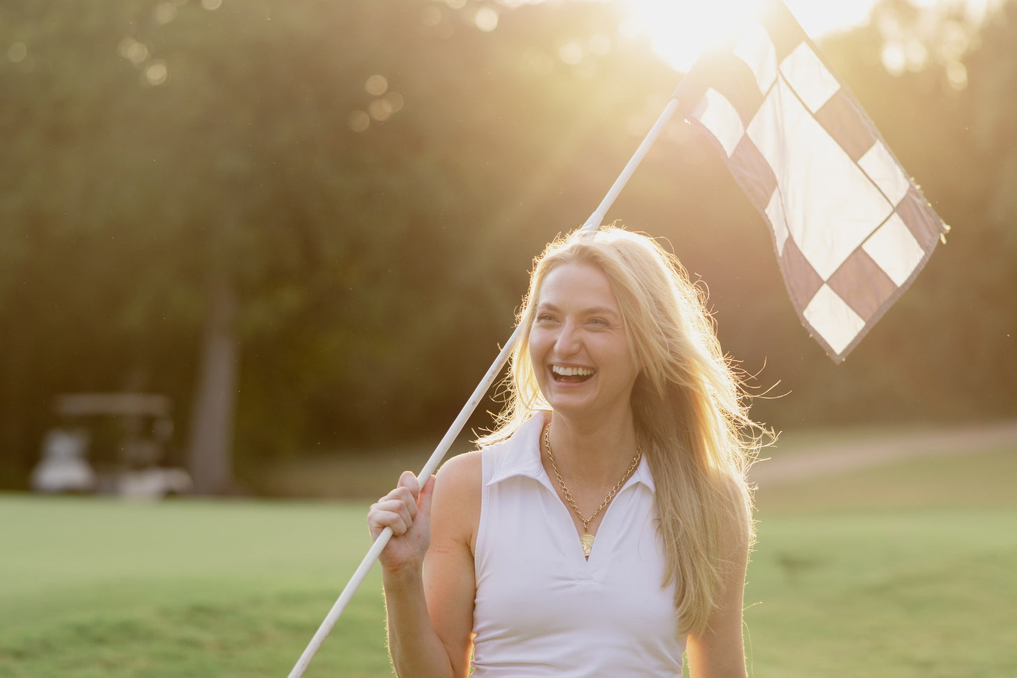 A woman carrying a checkered pin flag over her shoulder walks off a golf green in low evening light.